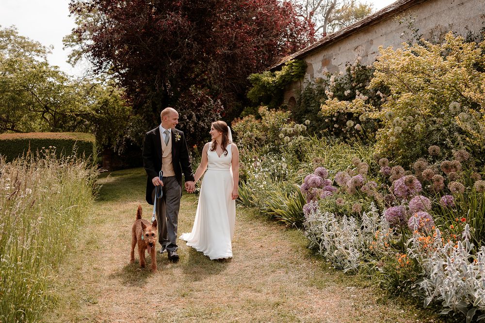 A couple walks their dog along a garden path with blooming flowers and stone walls.