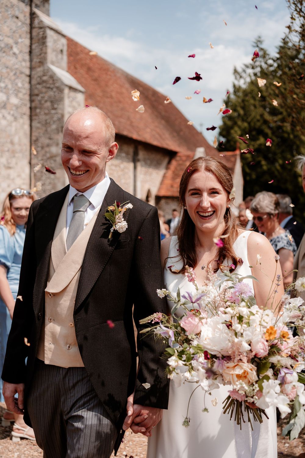 Newlyweds exit their wedding ceremony while guests throw flower petals in celebration outside a rustic stone building.