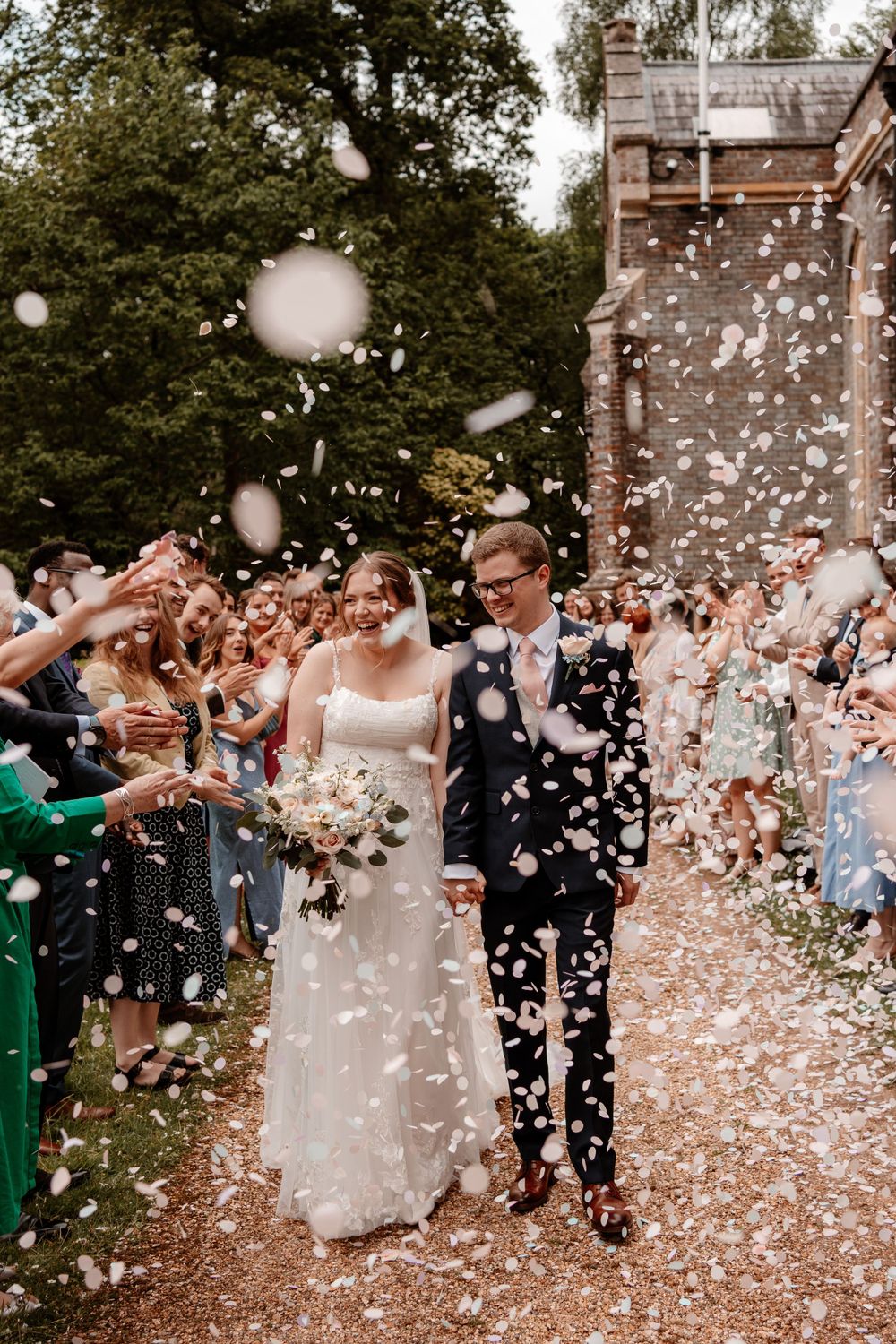 Wedding couple exits church while guests shower them with white confetti petals in joyful celebration.