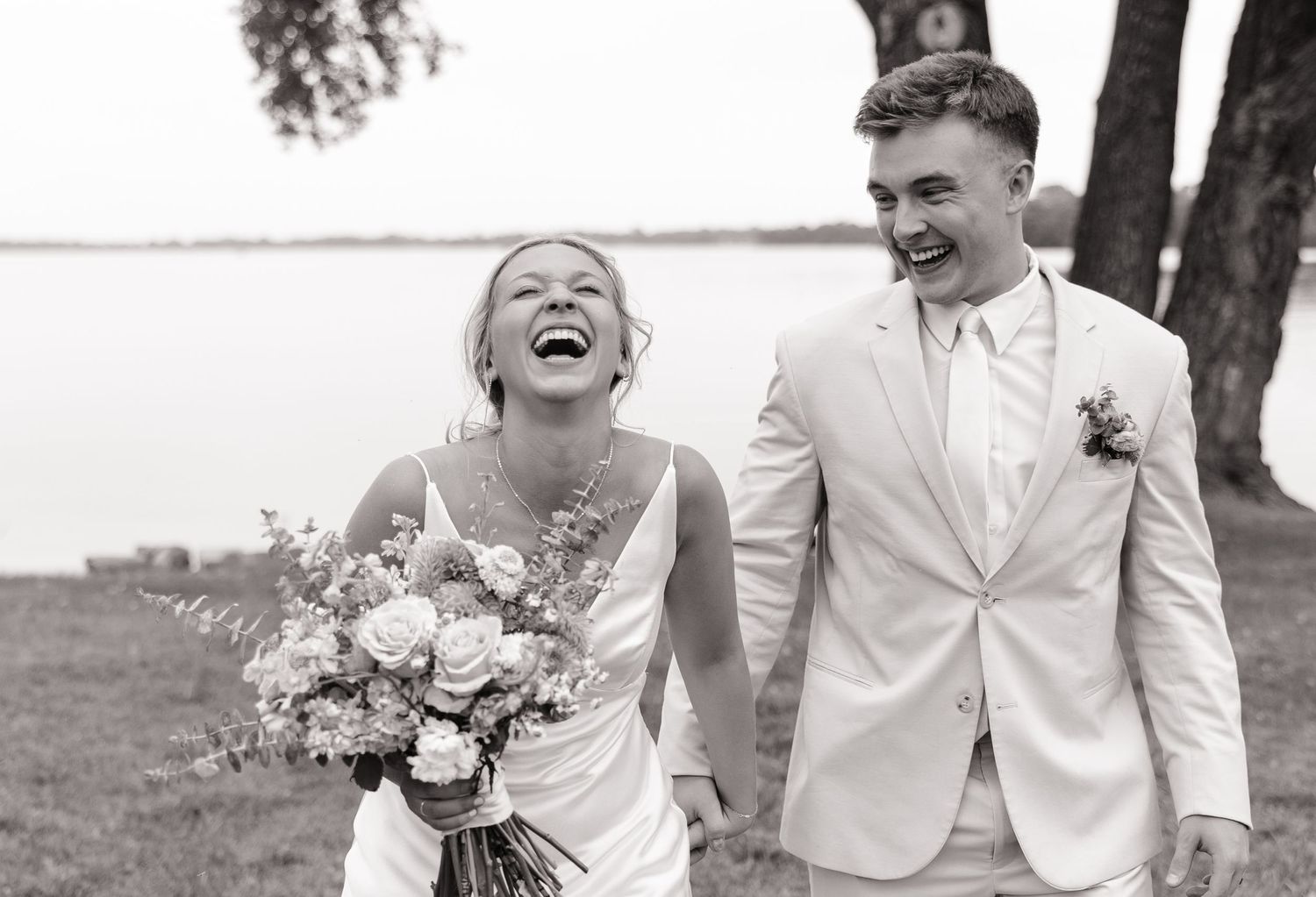 A bride and groom share a candid laughing moment on their wedding day by the water.