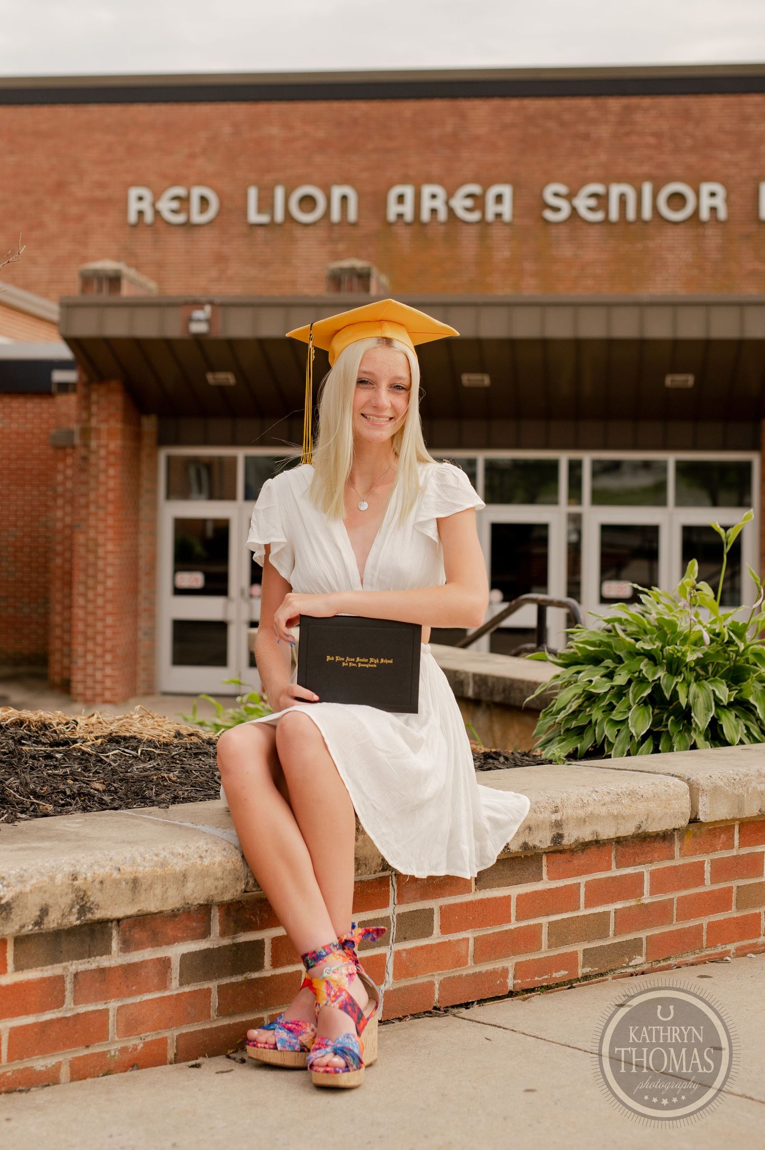Graduate in cap and gown sits on brick wall holding diploma at school entrance.