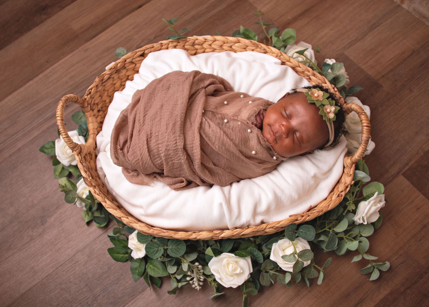 Newborn baby sleeping in brown wrap in floral basket on wooden floor.