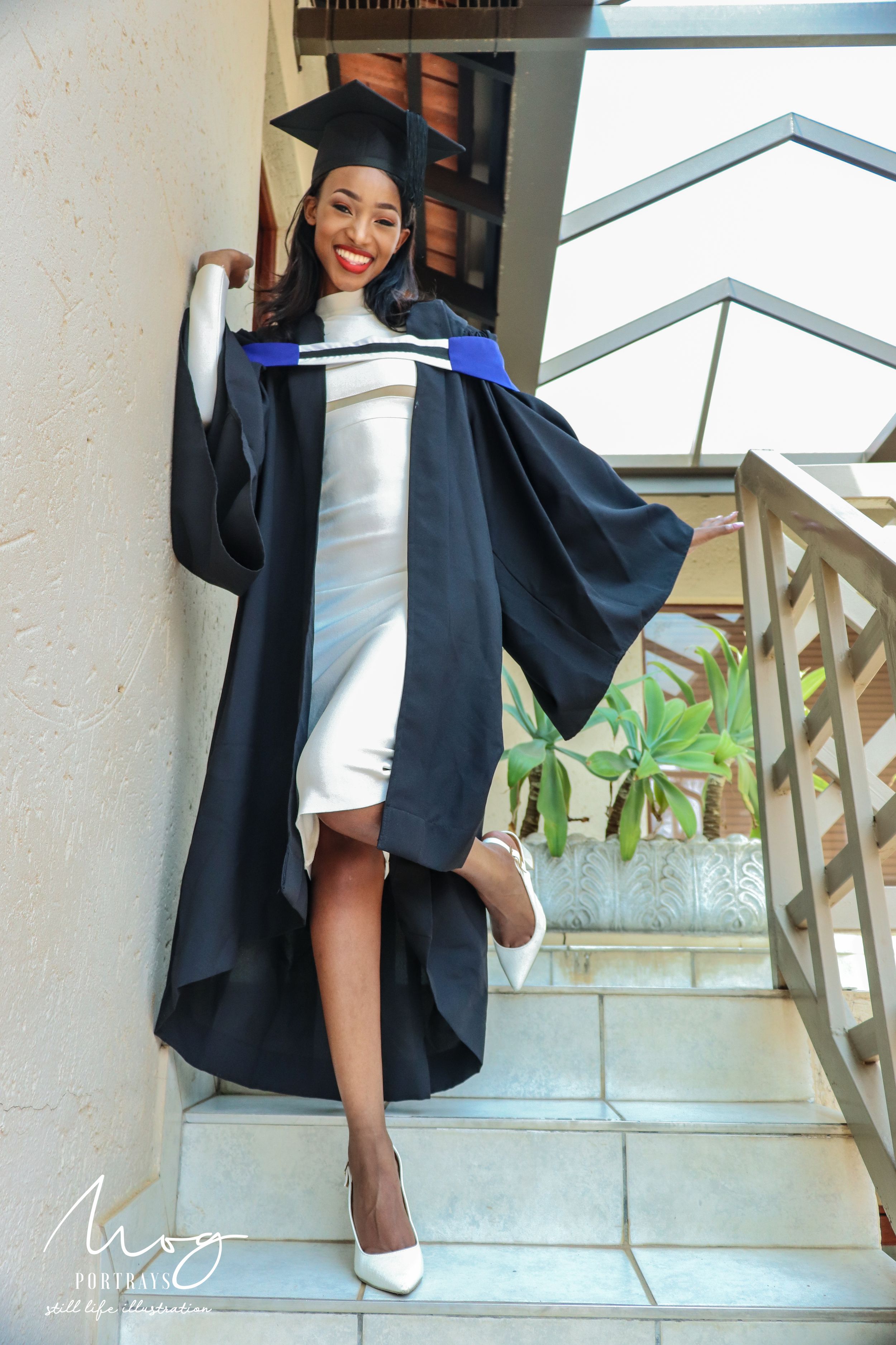 A graduate in white dress and black academic regalia poses joyfully on stairs with white heels and graduation cap.