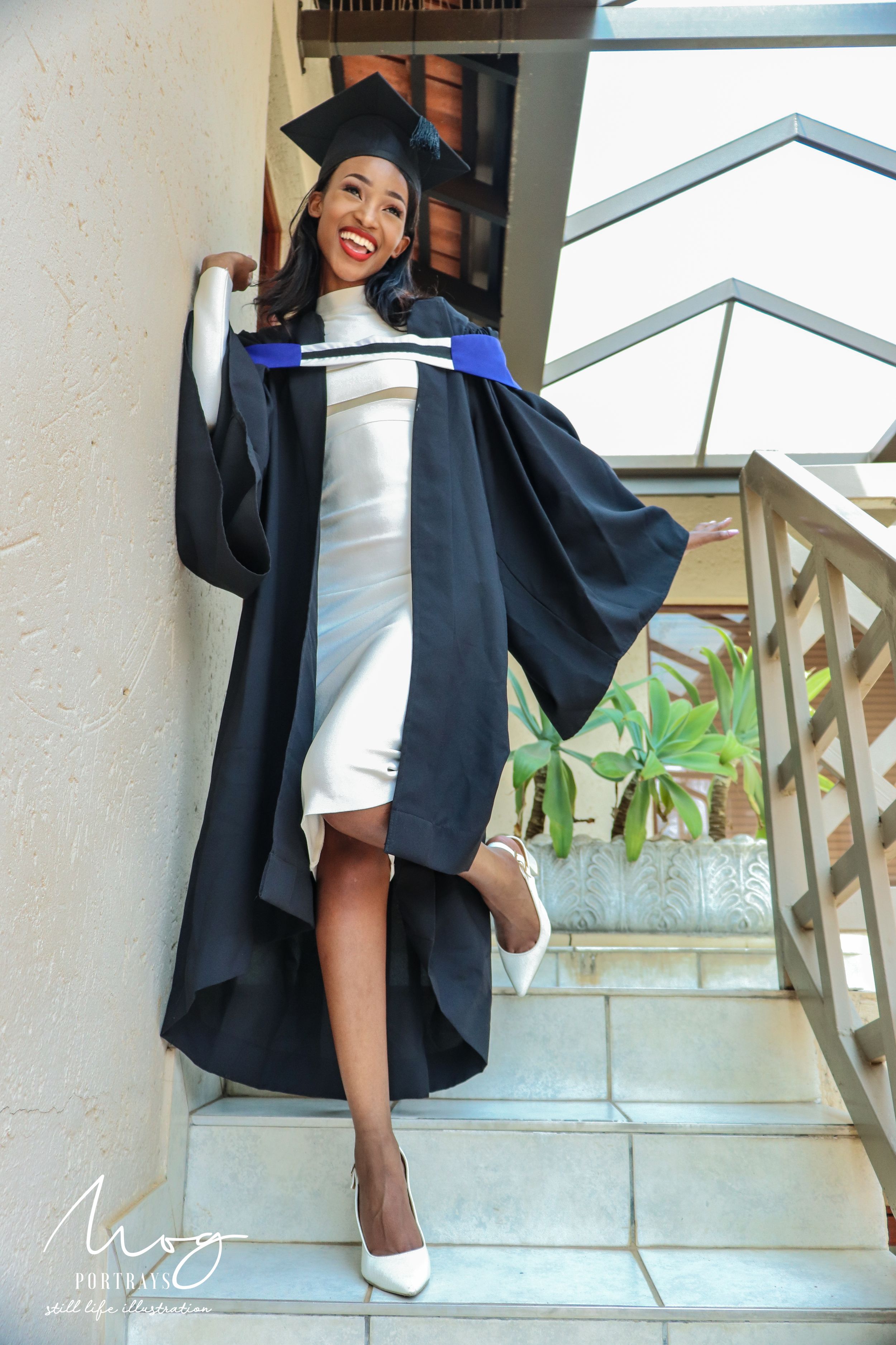 A graduate celebrates on stairs wearing white dress and black graduation gown while smiling brightly.
