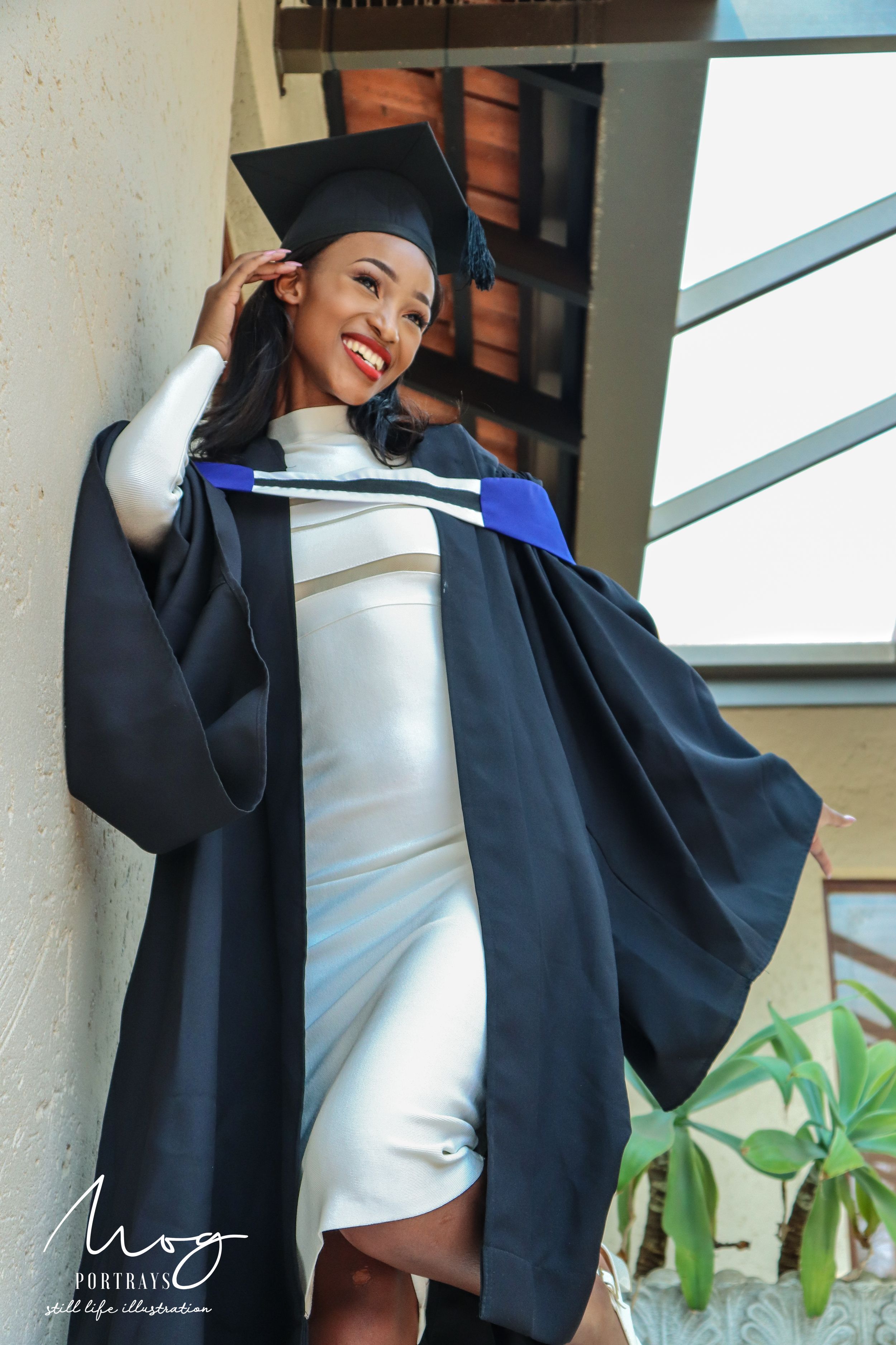 Smiling graduate in a white dress and black graduation gown poses by a window during a celebratory photoshoot.