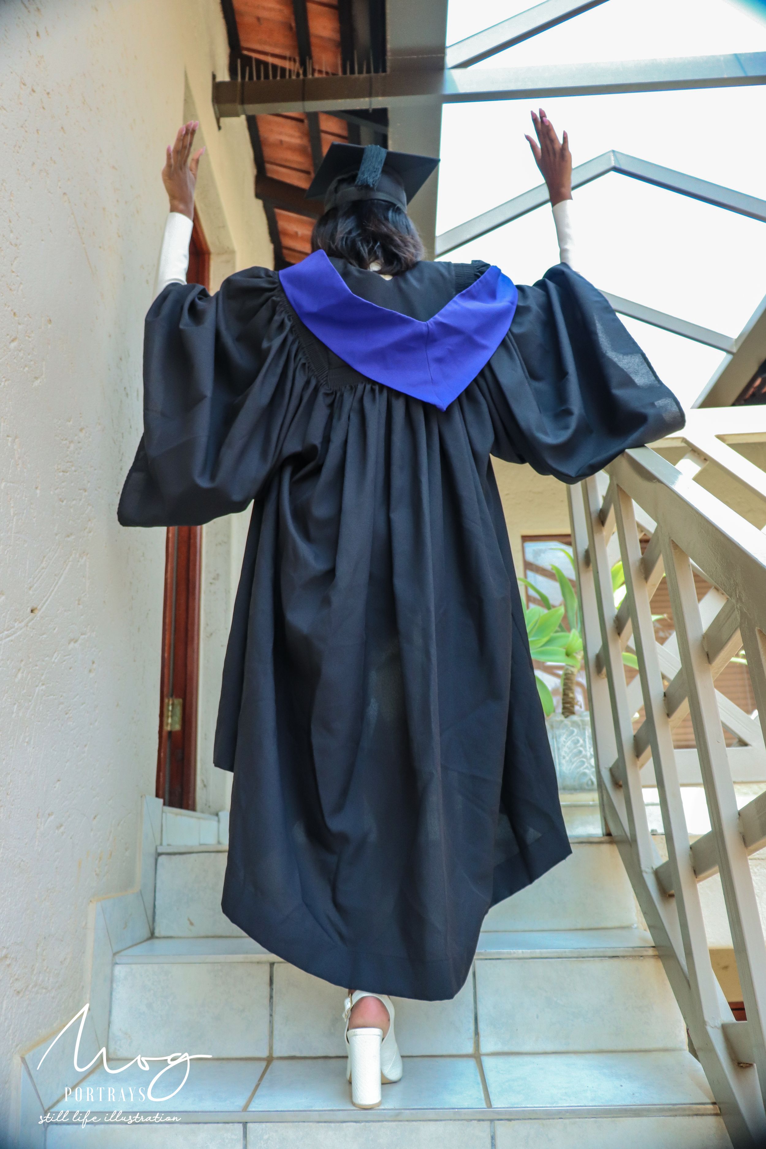Graduate in black cap and gown with blue stole raises hands in celebration on tiled steps.