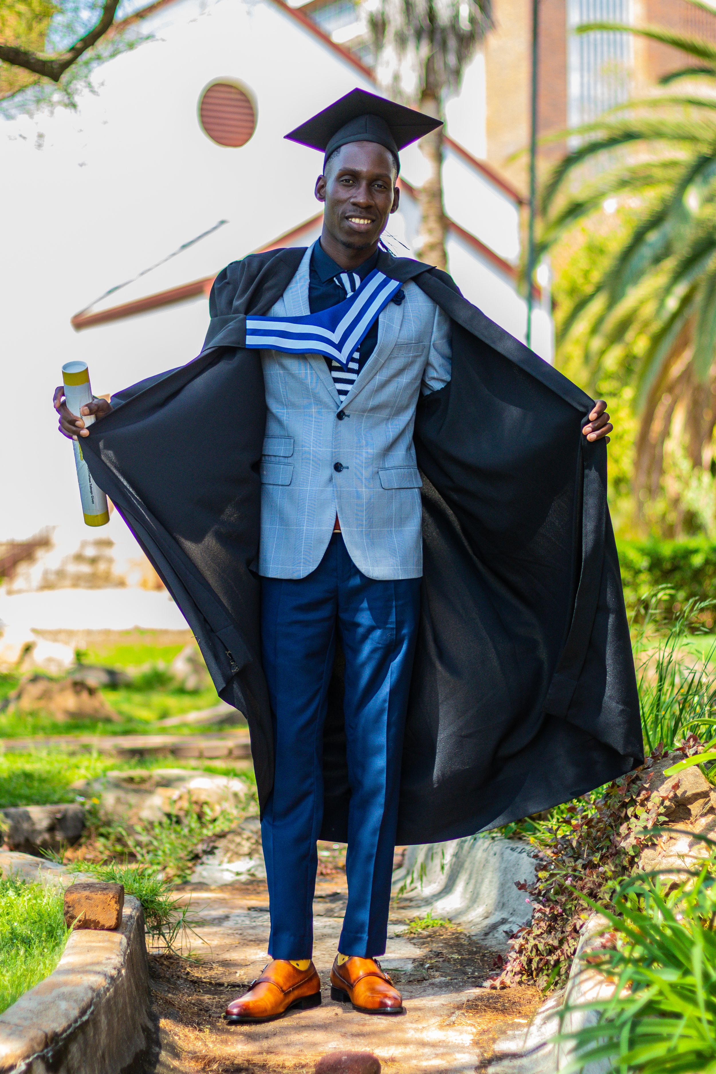 A graduate wearing a cap and gown poses outdoors showing off blue suit and brown shoes near tropical landscaping.