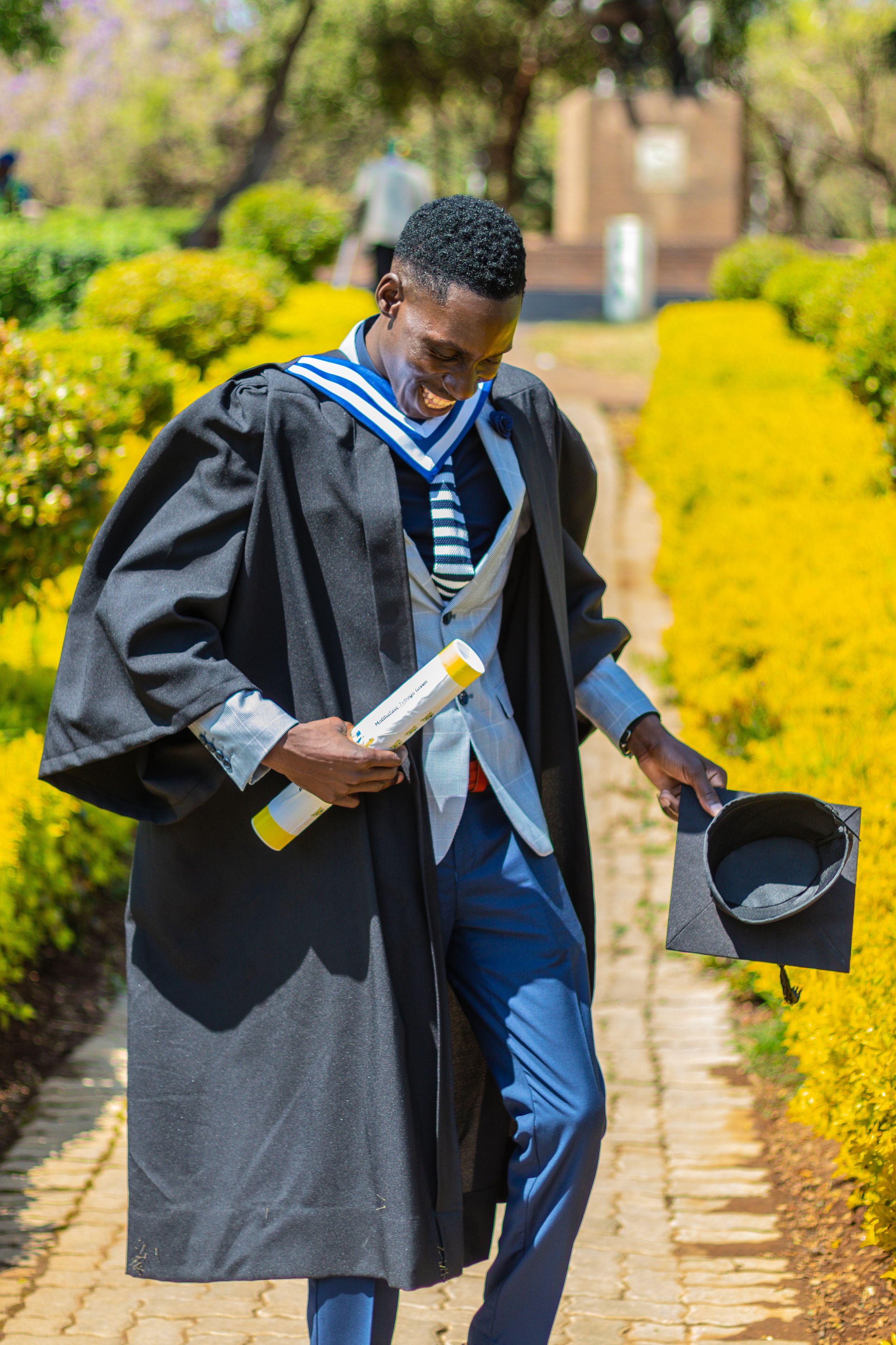 Graduate in academic regalia walks joyfully along garden path holding diploma and cap on sunny day.