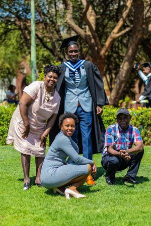 Group in formal attire gathered on sunny lawn during graduation celebration with green trees in background.