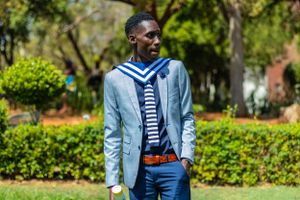 Young person in a light blue blazer and striped shirt poses outdoors in front of green landscaping on a sunny day.