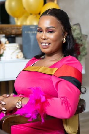 A person in a vibrant hot pink dress with gold accents smiles warmly while seated, with yellow balloons in the background.