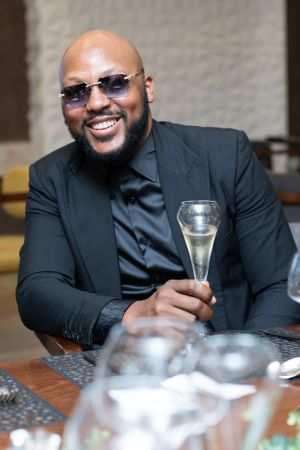 A well-dressed person in a black suit and sunglasses sits at a table holding a glass of champagne with a cheerful expression.