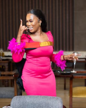 A smiling woman in a vibrant hot pink dress with feather details holds a champagne glass in an elegant setting.
