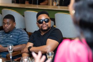 People enjoying drinks and conversation at an outdoor dining table with sunglasses and casual attire.