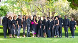 A group wearing black formal attire poses outdoors in a garden setting with green trees in the background.