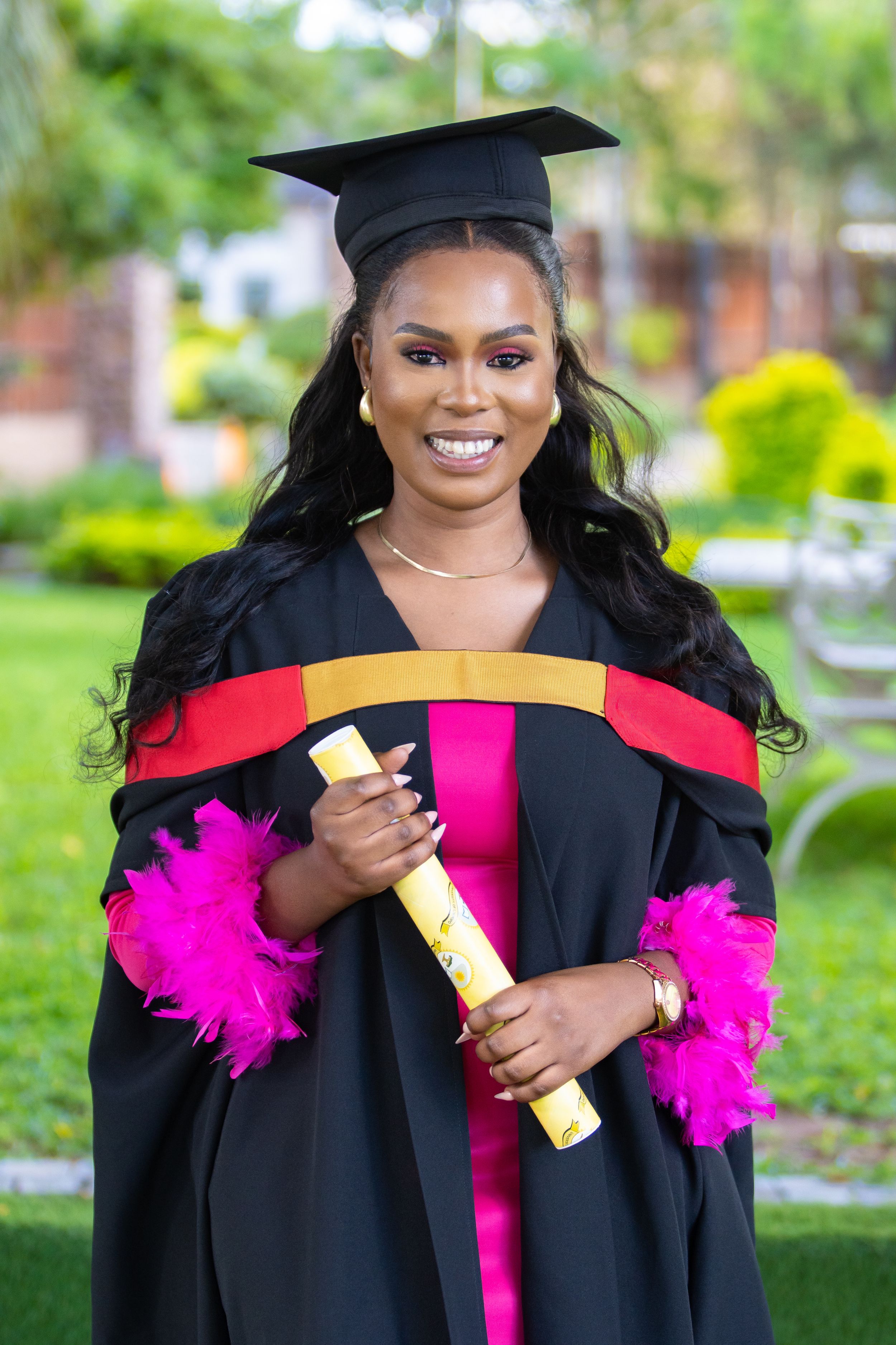 Graduate in academic regalia wearing black cap and gown with bright pink accents holds diploma during outdoor ceremony.