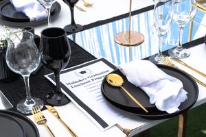 Elegant table setting with black plates, wine glasses, and gold utensils on a white tablecloth.