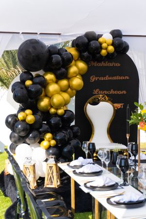 Elegant graduation party table setup with black and gold balloon arch, formal place settings and decorative backdrop.