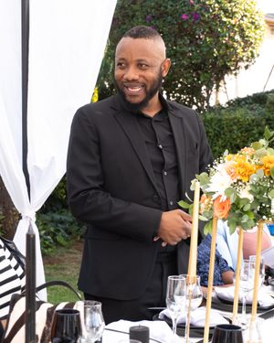 Person in a black blazer and dress shirt smiling at an elegant table setting with wine glasses and floral centerpieces.