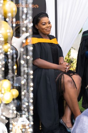 Graduate in black and yellow academic regalia poses at celebration with gold balloons and decorative backdrop.