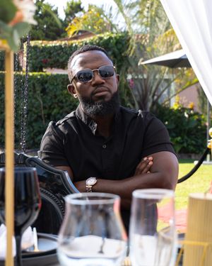 Someone wearing sunglasses and a black polo shirt sits at an outdoor table with wine glasses in the foreground.