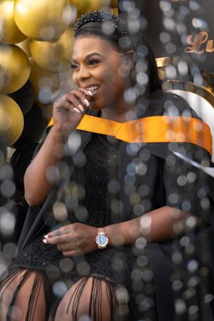 A graduation celebration with orange sash against a backdrop of gold balloons and sparkly bokeh lighting effects.