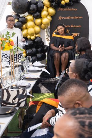 Elegant graduation party setup with black and gold balloon arch, formal table settings and guests seated at decorated table.