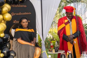 Academic graduation ceremony with two graduates wearing traditional caps and gowns alongside gold balloon decorations.