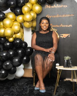A graduation celebration backdrop with black and gold balloon arch and decorative text on a dark wall.