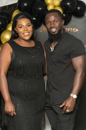 Two people pose together at an event with black and gold balloon decorations, wearing formal black attire.