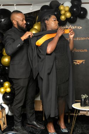 Two graduates in black regalia celebrate with balloons at a graduation party decorated with gold and black balloons.