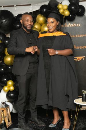 A graduation celebration with gold and black balloons decorating the backdrop as two people pose together in formal attire.