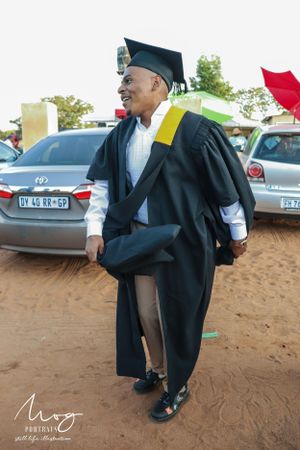 Graduate in black academic regalia with gold stole poses joyfully on campus grounds during graduation ceremony.
