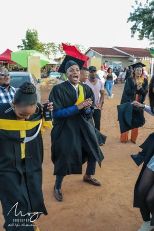 Graduate students in caps and gowns celebrate exuberantly on a dirt path during commencement.