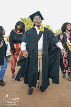 A graduate in cap and gown celebrates joyfully during commencement ceremony on a sunny outdoor day.