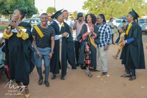 A group of graduates in black gowns celebrate together outdoors on a sunny day.
