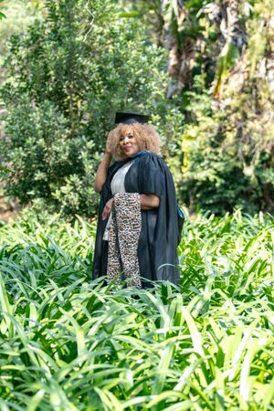 Graduate wearing a black cap and gown with leopard print leggings poses in a lush green garden setting.