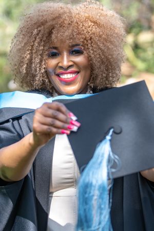 A graduate in academic regalia displays their diploma while smiling outdoors in natural lighting.