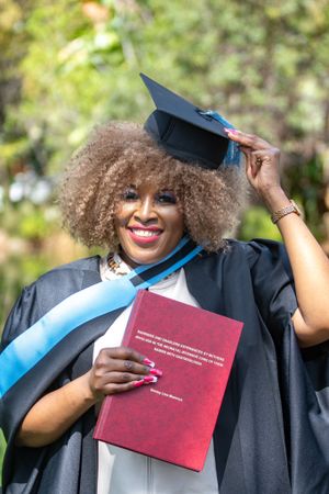Graduate in academic regalia holds their thesis or dissertation while adjusting their mortarboard cap with natural curly hair.