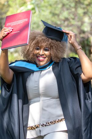 Graduate in black academic regalia and natural curly hair celebrates with diploma and cap during graduation ceremony.