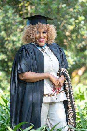 Graduate in traditional black cap and gown poses outdoors holding leopard print accessory.