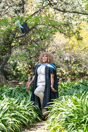 Graduate tosses academic cap into the air while celebrating on garden path surrounded by lush greenery.
