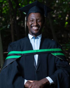 Graduate in black cap and green-trimmed academic regalia smiles brightly during graduation ceremony outdoors.