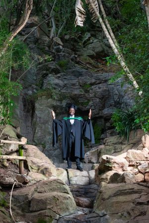 A graduate in academic regalia stands on rocky steps surrounded by forest and stone walls in nature.