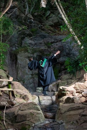 A graduate in black cap and gown stands triumphantly on rocky terrain surrounded by trees and natural stone formations.