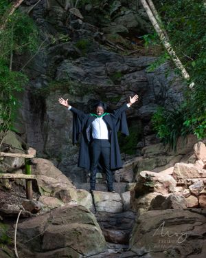 A person in formal attire and black cape poses dramatically with arms raised on rocky terrain in front of a cave opening.