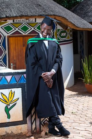 A graduate in academic regalia stands outside a traditional African-style building with painted geometric patterns and thatched roof.