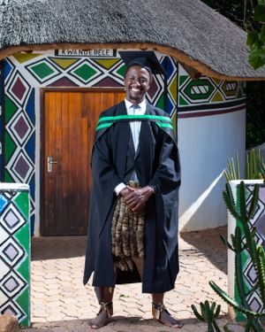 Graduate in traditional academic regalia poses in front of a thatched-roof building with colorful geometric Ndebele patterns.