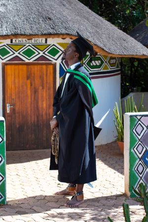 Graduate stands proudly in academic regalia outside a traditional African-style building with geometric patterns.