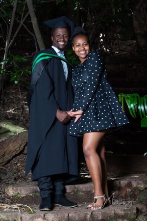 A graduation photo featuring a graduate in cap and gown with green sash standing next to someone in a polka dot dress outdoors.